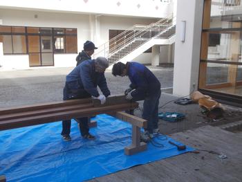 中庭のデッキに敷かれたブルーシートの上で、複数の作業員が木材の加工や組み立てを行っている様子の写真