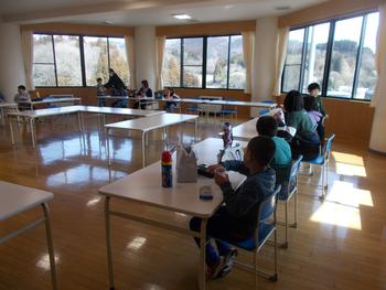 机がコの字型に配置された広い部屋で、小学生たちが食事や休憩をとっている様子の写真