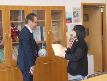 書類棚が置かれた室内で、スーツを着た男性と黒い上着の女子児童が向かい合って立ち、女子児童が手に持った茶色の封筒を手渡そうとしている様子の写真