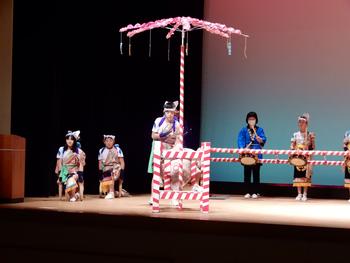 ステージ上で伝統的な衣装を着た人々が、男性が持つ大きな傘と縞模様の柵を使いながら踊りを披露しており、後ろでは太鼓を叩く人もいる様子の写真