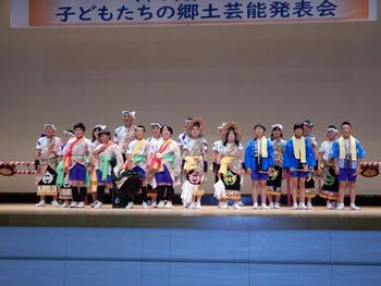 ステージ上に和服やカラフルな伝統衣装を身につけた大勢の子どもたちが横一列に並んでおり、背景には「子どもたちの郷土芸能発表会」と書かれた横断幕がある写真