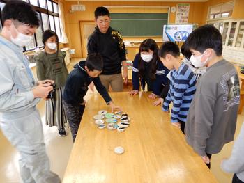 教室で児童たちがテーブルの上に並べられた作品を囲んで興味深そうに見ている様子の写真