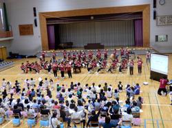 体育館の舞台の下に竹崎高級中学校の56名の生徒が楽器を演奏し児童達が並べられた椅子に座り演奏を聞いている写真