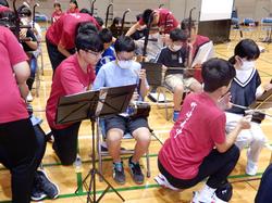 竹崎高級中学校の生徒が演奏していた場所に児童が楽器を持ち座り、その横に竹崎高級中学校の生徒がしゃがんで楽器の弾き方を教えている写真