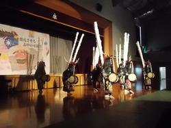 伝統的な装束を身につけ、太鼓を抱えた複数の人物が、舞台の前で演舞を披露している写真