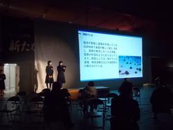 照明を落としたステージで、2人の生徒がスクリーンに映し出されたスライドを使って発表を行っている写真