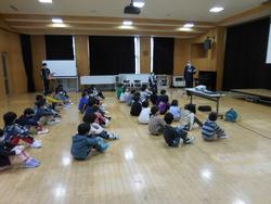 前方にスクリーンや机が並んでいる広い室内のホールで子どもたちが床に座って前方の所長さんの話を聞いている写真