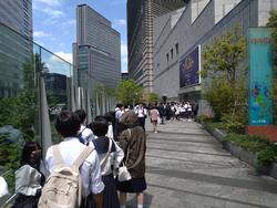 高層ビルの立ち並ぶ街中の歩道で、多くの制服姿の生徒たちが列を作って建物の入口へ向かって歩いている写真