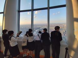 東京スカイツリーの展望台の大きな窓から外の夕景を眺めている生徒たちの写真