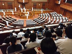 木製の座席と机が半円形に並ぶ国会議事堂の本会議場を、制服姿の生徒たちが傍聴席から見学している写真