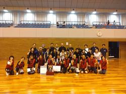 スポーツ大会の会場で、ユニフォームを着た複数の選手たちが優勝トロフィーや賞状を囲んで集合写真を撮っている写真