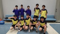 水色の長椅子が置かれた室内で、黄色と黒のユニフォームや学校のジャージを着た生徒たちが並んでピースサインをして写した記念写真