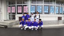 窓に「百花繚乱」などの文字が書かれた紙が貼られた校舎の窓の前で、野球部のユニフォームを着た男子生徒たちが並んで写した記念写真