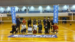 体育館のバレーボールコートで、2列に並んだ志津川Bチームメンバーの前列中央の2人が賞状とトロフィーを持ち、後列両端の2人が青い旗を持って、横断幕の前で記念撮影をしている写真