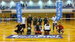 体育館のバレーボールコートで、2列に並んだ志津川Cチームメンバーの前列中央の2人が賞状とトロフィーを持ち、後列両端の2人が青い旗を持って、床に置かれた横断幕の前で記念撮影をしている写真