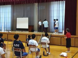 壇上にプロジェクタースクリーンが置かれ、その横で講師と数名の生徒が輪になって立っている様子を、下に並べた椅子に座った生徒たちが見ている写真