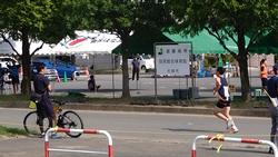 白や緑のテントが街路樹の後ろに並び、その前の舗装された道路を、ユニフォームを着てゼッケンをつけた男子生徒が走っており、沿道では男性が自転車を降りてその様子を見ている駅伝大会の写真