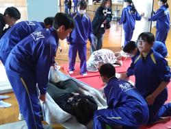 体育館で、体操服姿の生徒たちが毛布を担架代わりに使い、負傷者役の生徒を運ぶ救護訓練を行っている写真