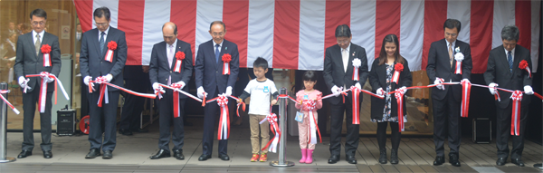 黒いスーツ姿の男性たちと子どもたちが、赤と白の紅白幕を背景に、白い手袋をはめて紅白のリボンを切っている開業イベントの様子の写真