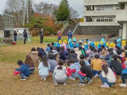 校庭に多くの児童が集まり座って前方でマイクを持って話している方の話を聞いている写真