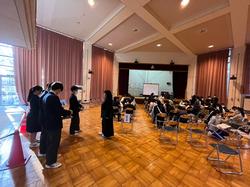 ホールの端に数人の生徒が並んで立ち、その奥には大勢の生徒や参加者たちが椅子に座り見学会に参加している様子の写真