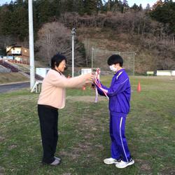 芝生の広場でジャージ姿の男子生徒が女性から優勝トロフィーを受け取っている写真