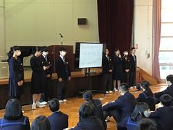 舞台の前に置かれた大きなモニターの両側に制服姿の生徒が4人ずつ立ち、前を向いて床に座った生徒たちに話をしている写真