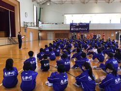 体育館に集まった大勢の生徒たちが、舞台前に立つ先生の話を聞きながら静かに座っている集会の写真