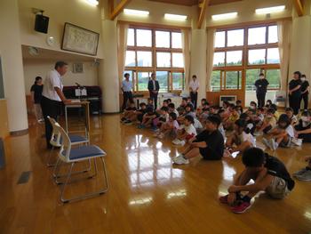 広い講堂に集まったたくさんの児童が床に整列して座っており、後方に立っている教職員たちと一緒に、前方の教卓に立った校長先生の話を聞いている写真