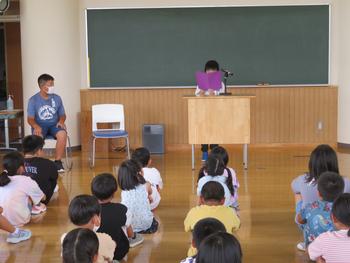 一人の児童が教卓に立ち、マイクを使って紫色のノート見ながら発表し、他の児童たちは床に座って、前を向いてしっかり聞いている始業式の様子の写真