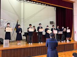 ステージの上に賞状などを持った生徒たちが横一列にずらっと並び、ステージ下に一人の女子生徒が座っている写真