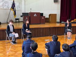 体育館のステージの前に置いた3脚の椅子にそれぞれ女性が座り、右側の女性がマイクで話をしている様子を体育座りをした生徒たちが見ている写真