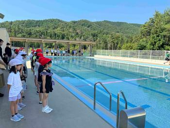 白い帽子や赤い帽子をかぶった小学生たちがプールサイドに一列に並び、関係者が見守る中、水泳の授業が行われている様子の写真