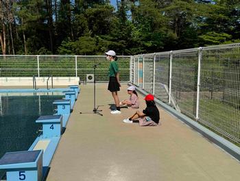 プールサイドで白帽をかぶった児童がマイクの前に立ち、他の児童2人が横で座って見守る中、開会のあいさつをしている様子の写真