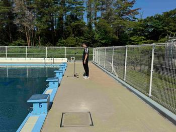 屋外プールサイドで、マイクスタンドの前に立ち、プールに向かって話している男性の写真