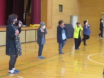 体育館のステージ前で数人の大人が並び、体操のような動きをしながら話している写真