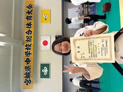 「宮城県中学校総合体育大会」と書かれた横断幕が掲げられた会場で、柔道着を着て賞状を片手にピースサインをしている芳賀さんの写真