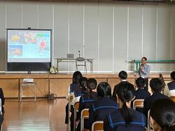 体育館のステージ上に設置されたスクリーンに資料が映し出され、席に座った生徒たちがステージ前に立った男性の講話を聴いている様子の写真