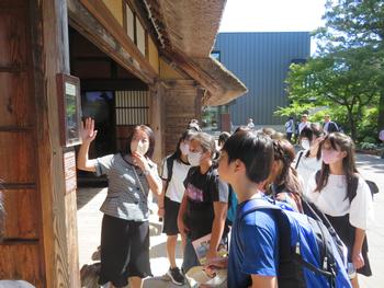 伝統的な日本家屋の前で、マスクをした女性ガイドが、周りに集まった児童たちに展示物の説明をしている様子の写真