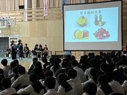 体育館の壁に設置された大きなスクリーンに果物の写真が映し出され、台湾の竹崎高級中学校の発表が行われており、大勢の生徒たちが座ってそれを見ている写真