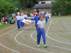 先頭は半袖の体操服と青いジャージのズボンを着た男子児童、その後ろには日の丸の旗を横に持っている児童たちが運動会の入場行進をしている写真