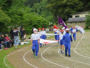 運動会で国旗を持った児童を先頭に旗などを掲げた体操着姿の児童たちがトラック内を入場行進をしている写真