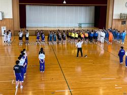 体育館に集まった部活のユニフォームを着た生徒たちが、部活動ごとに整列し、向かい合って中央に立った男性が右手を上に挙げている壮行会の様子の写真