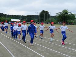 屋外のグラウンドで、赤い帽子をかぶった児童たちが整列して行進している写真