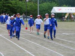 先頭の青いジャージ姿の児童がいて、手を大きく振りながら真剣な表情で行進し赤白帽をかぶった児童たちが、列をなして前方へ歩いている運動会の入場行進の写真