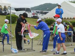 青いジャージを着た1人の児童が、トロフィーに飾るたくさんの赤白のリボンを受け取っている運動会の表彰の場面の写真