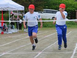 2人の男子児童が赤い帽子をかぶり、白いTシャツと青いズボン姿でゴールテープに向かって全力で走っている写真