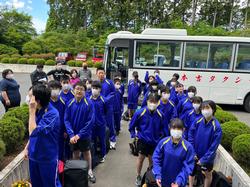 それぞれ荷物を持った青いジャージを着た多くの生徒たちが白いマイクロバスの前に集まり、整列している様子の写真