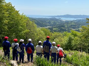 山の展望台から赤白帽子と青い体操服姿で、海や山が広がる雄大な景色を眺める小学生たちの後ろ姿が写された写真