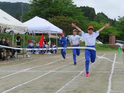 3人の児童が全力でゴールに向かって走り、手前の児童は赤い帽子をかぶり、両手を大きく広げながら笑顔でゴールテープを切っている様子を写した写真